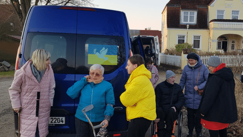 Mehrere Personen stehen vor einem Kleinbus, ein Mann sitzt im Rollstuhl, zwei Frauen haben Gehhilfen dabei.